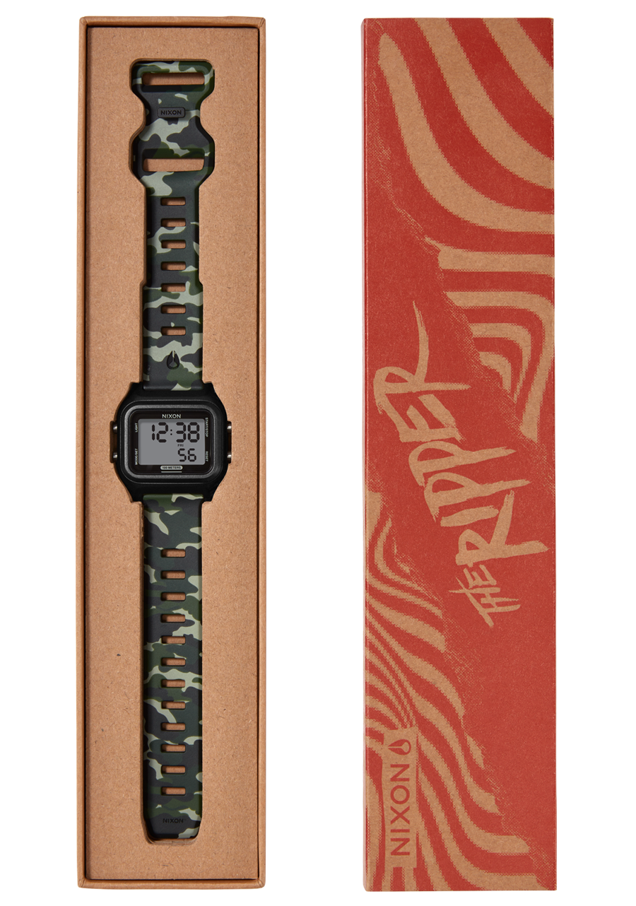 A Nixon "The Ripper" digital watch with a camouflage band is displayed in an open brown cardboard box next to its red and tan patterned sleeve.