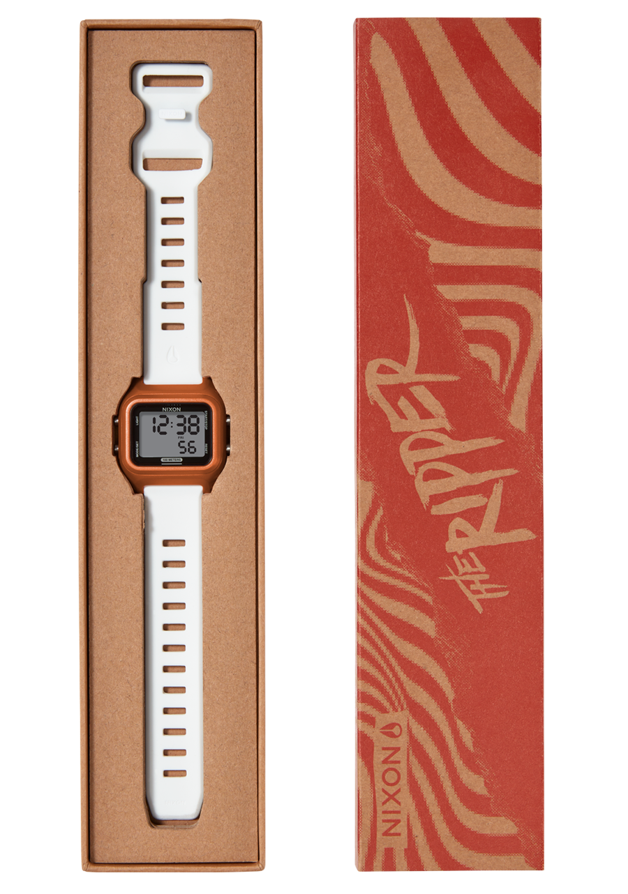 A Nixon "The Ripper" watch with an orange face and white band is displayed in a brown cardboard box next to its red and brown patterned sleeve.