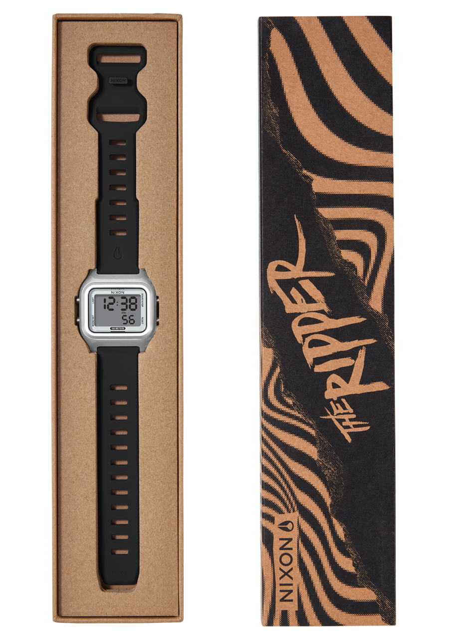 A silver digital watch with a black strap is displayed in a brown cardboard box next to its black and brown striped packaging that reads "THE RIPPER" and "NIXON".