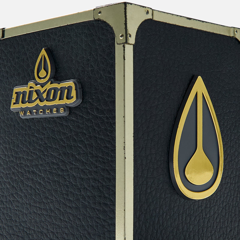 Close-up of a black textured case with gold corner protectors and two Nixon Watches logos, one with text and one with a teardrop symbol.