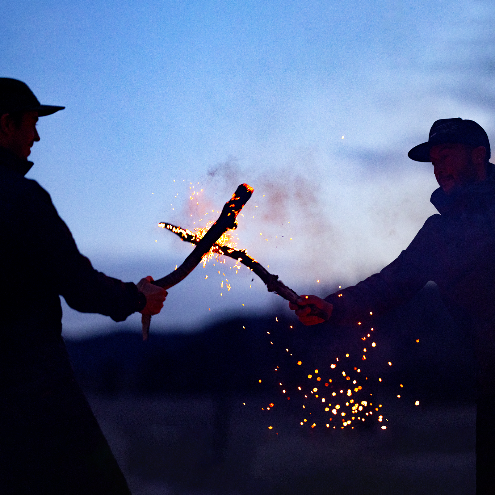 Two men play with fire logs while wearing Nixon gear outside at night.